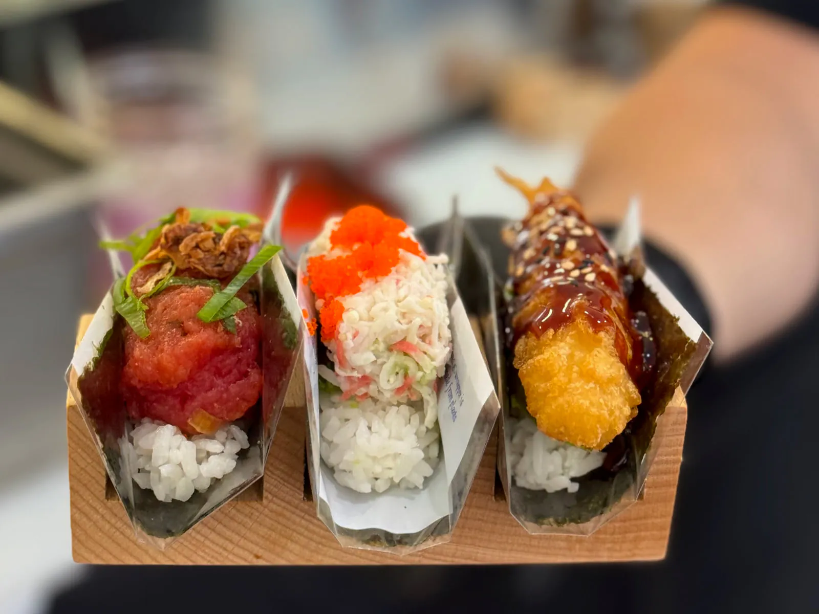 Three hand rolls in a wooden holder, held out across the counter