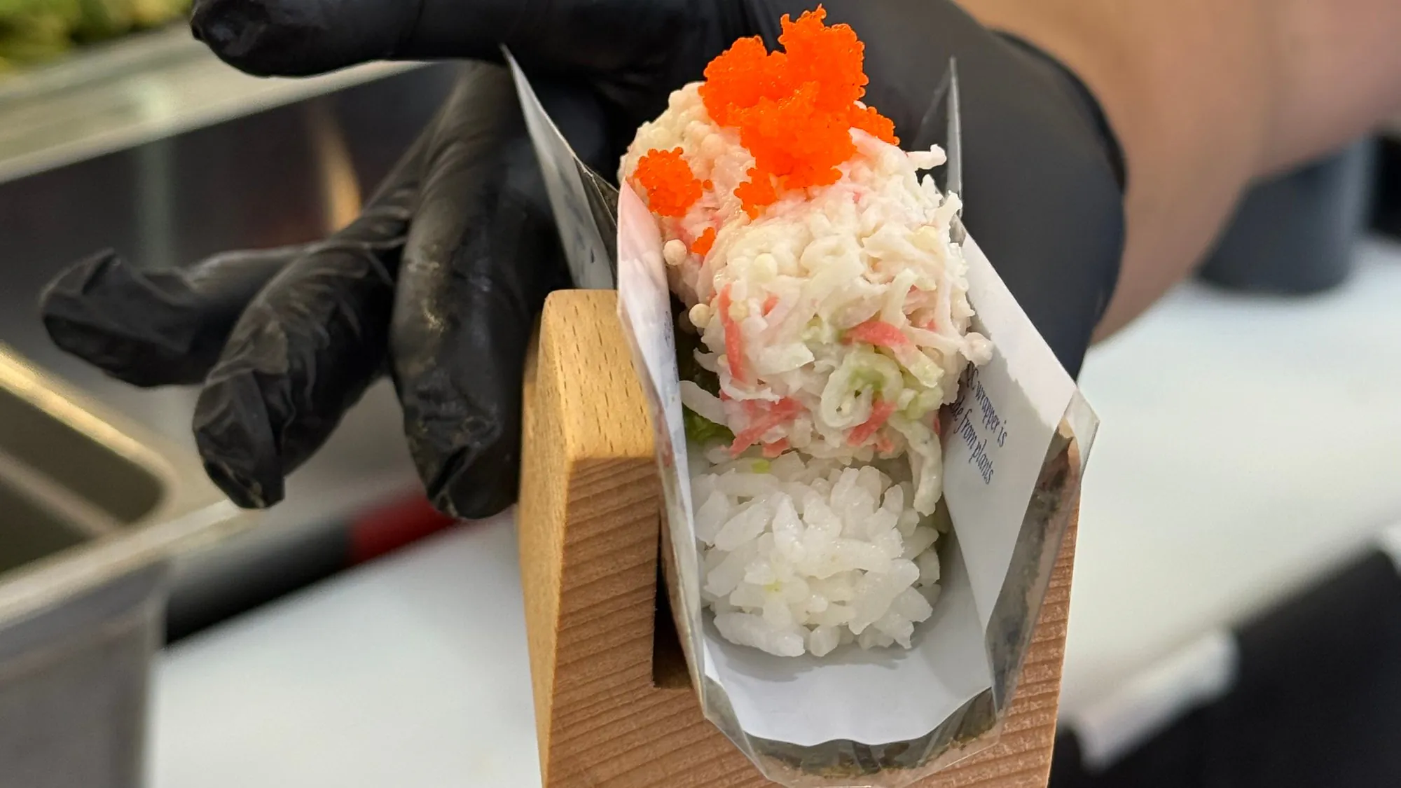 A chef in a black glove holding out a freshly rolled hand roll in its wooden stand