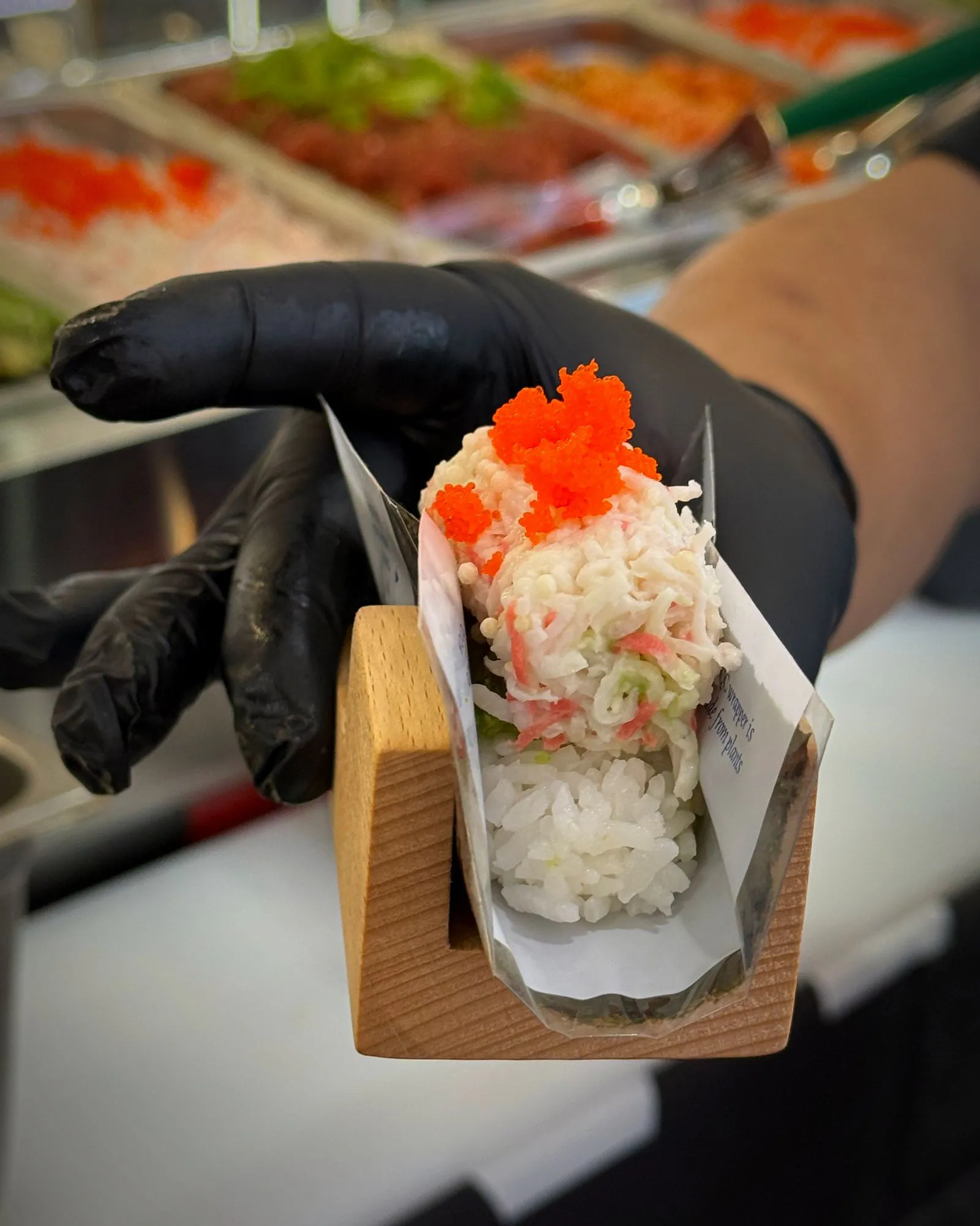 A chef in a black glove holding out a California hand roll in its wooden stand