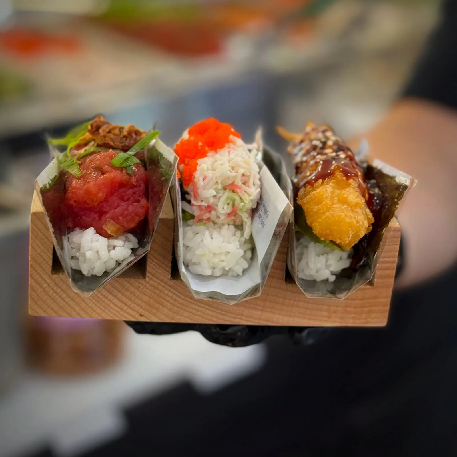 Three hand rolls in their wraps in one wooden holder — spicy tuna, California and shrimp tempura — held out across the counter